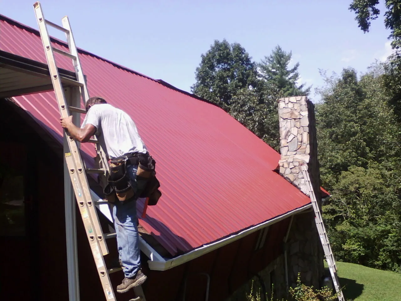 Roofer on a ladder beside a red metal roof and stone chimney — Soffit & Fascia Repair by Roofing Brothers in Pocono Lake